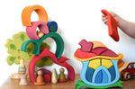 A child playing with the Glückskäfer Mushroom House wooden stacking toy. They have taken the red roof piece off the stack. The red gable house toy can be seen in the background  staked up to create a tower. 