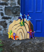 The Glückskäfer Natural Grotto placed on a stone wall in front of a blue door. Magic Wood felt elves have been placed on the pieces of the grotto. Some are in a seating position whilst others are standing.