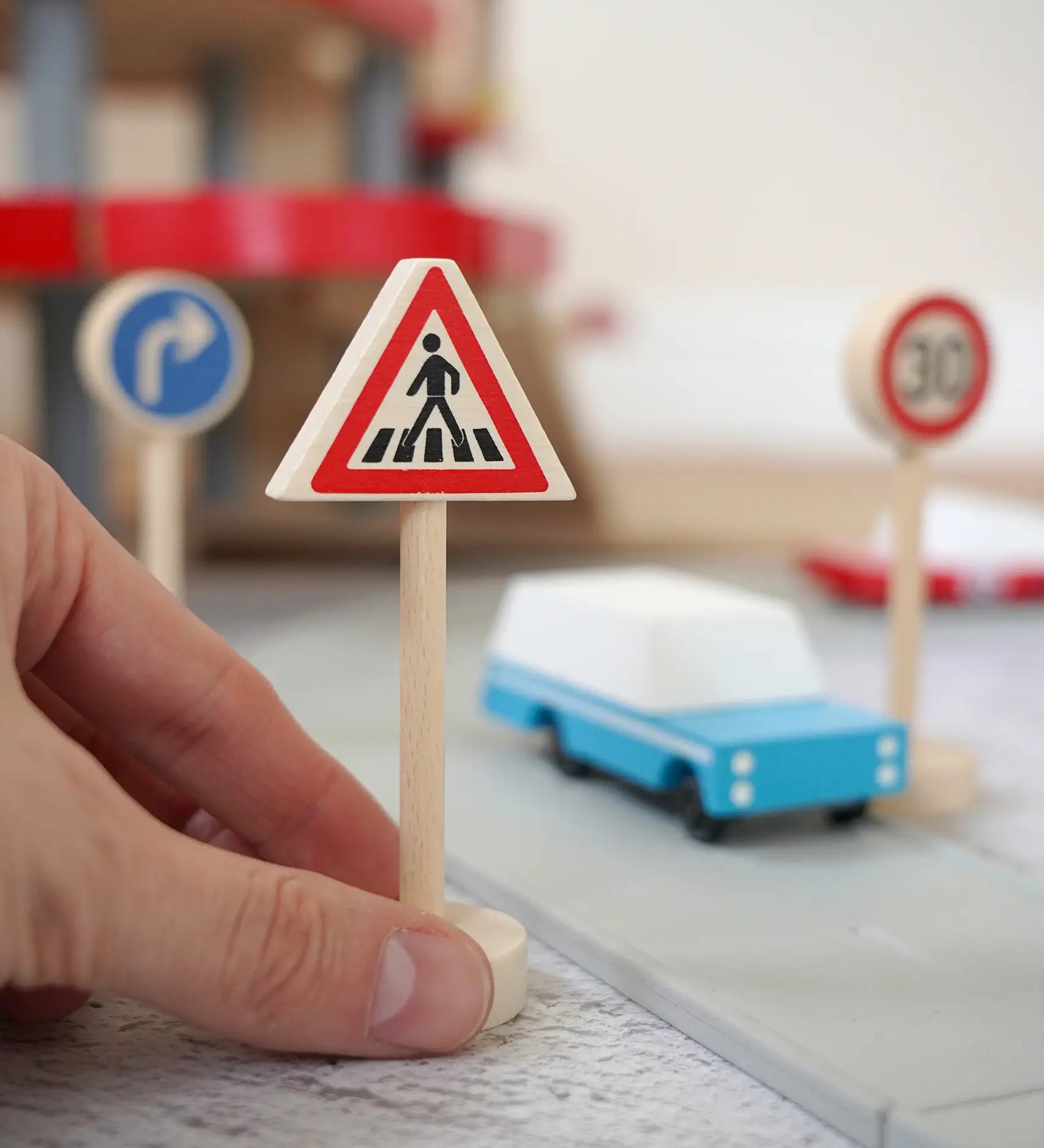 Adult hand holding the Gluckskafer miniature zebra crossing traffic sign next o a play road set