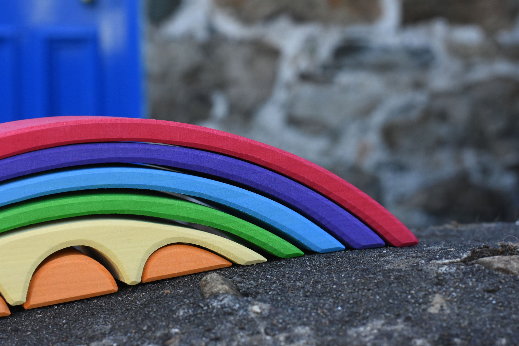 A close up of the front of the Gluckskafer Rainbow Bridge wooden toy. The bridge has been placed on a stone wall outdoors. 