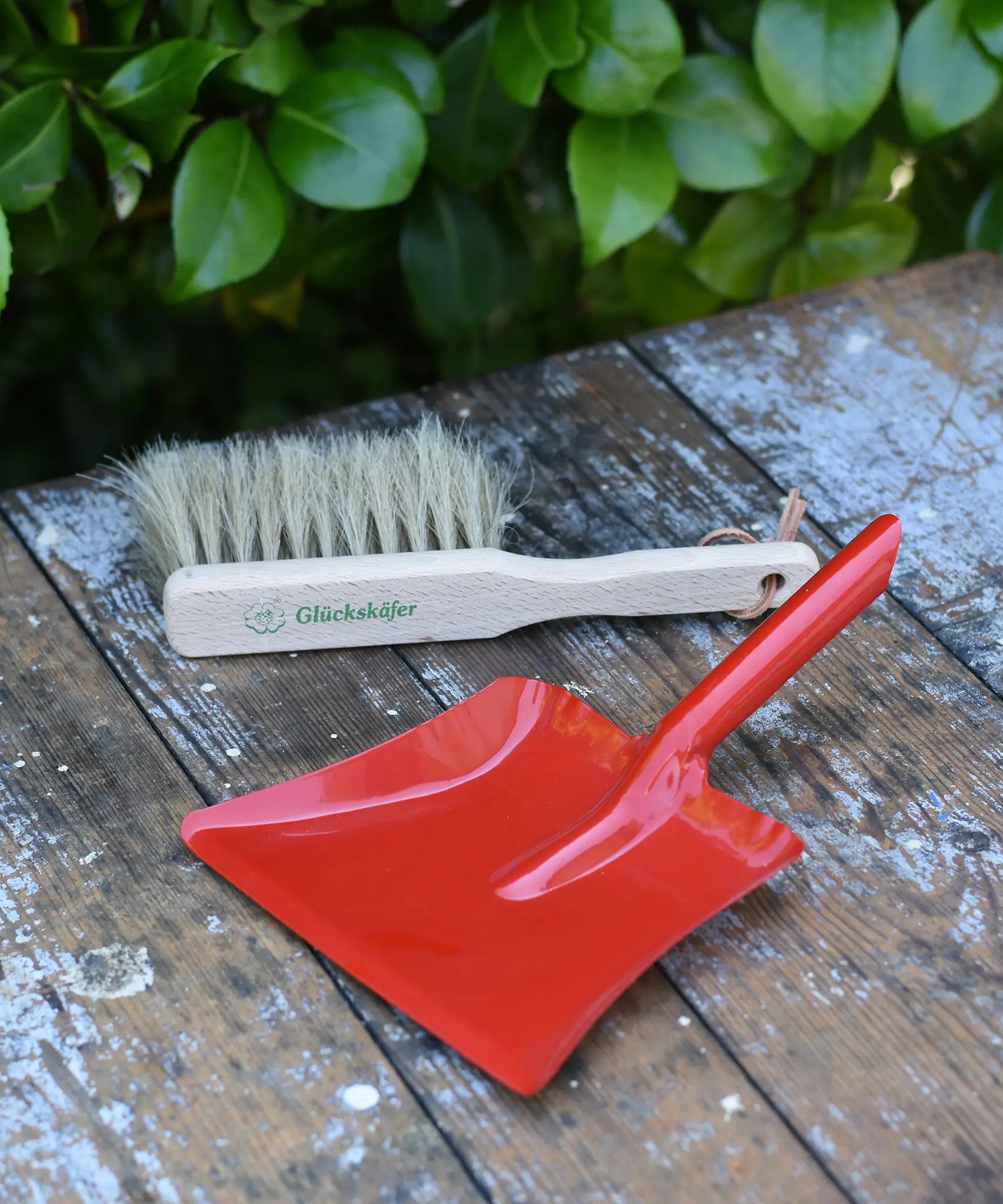 Glückskäfer red metal dust pan and dust brush placed on a wooden surface.