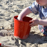 A young child holding the Glückskäfer red Metal Bucket, that has been placed on sand a a beach.