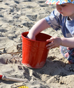 A young child holding the Glückskäfer red Metal Bucket, that has been placed on sand a a beach.