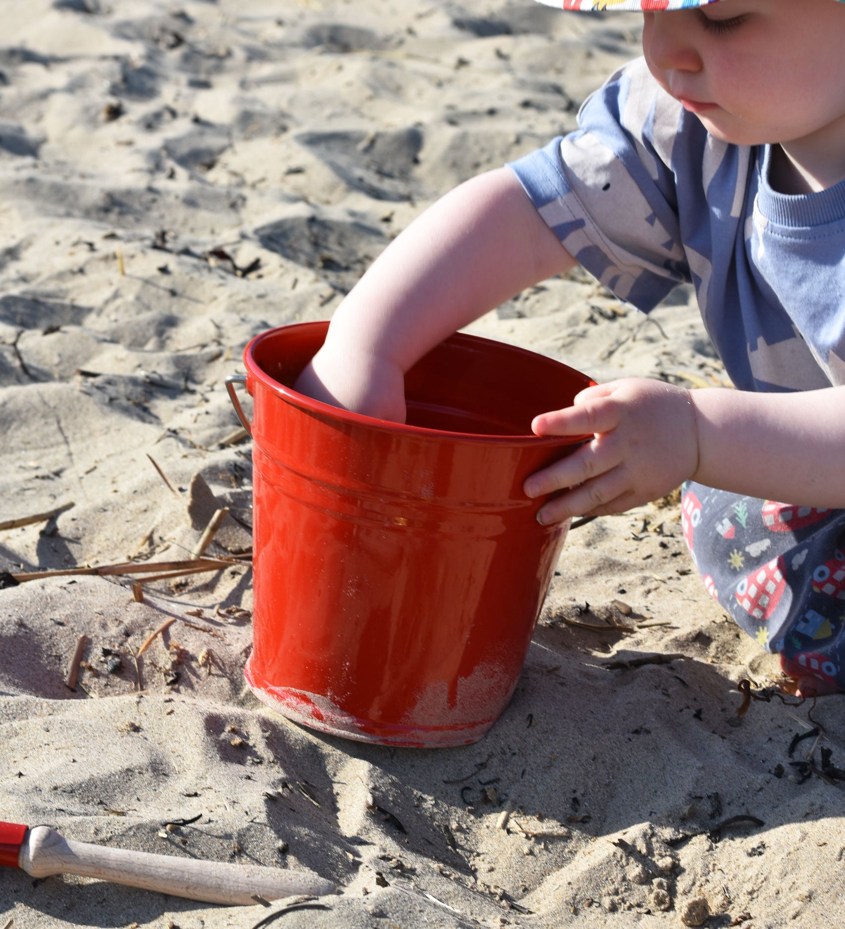 A young child holding the Glückskäfer red Metal Bucket, that has been placed on sand a a beach.