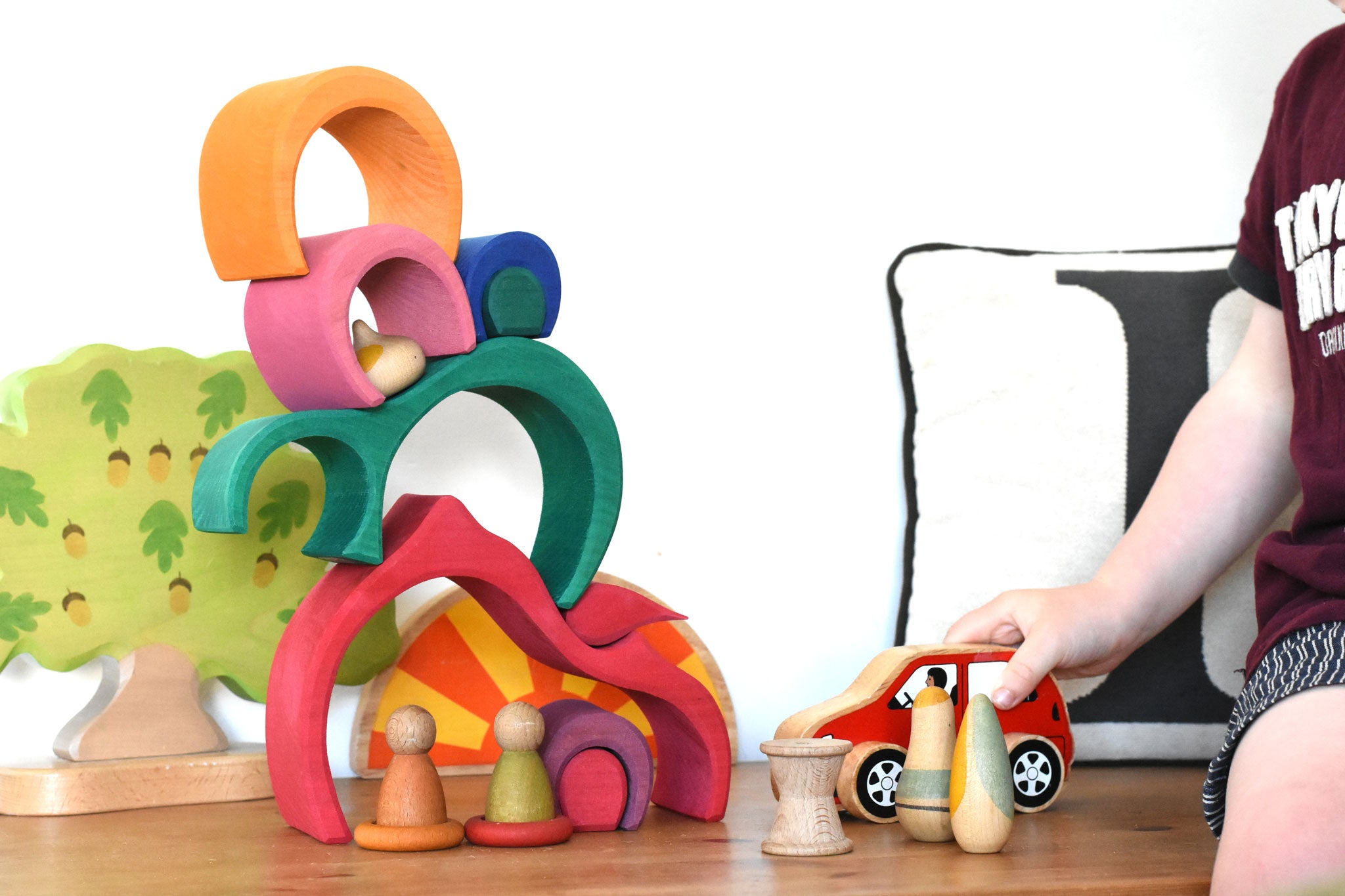 A child playing with a red Lanka Kade push along car with a Glückskäfer Gable House wooden stacking toy, stacked up in the background. Other wooden toys such as the Grapat Nins, wooden spools and a Holztiger oak tree have been added to the small world play scene set up. 