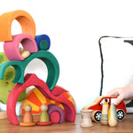A child playing with a red Lanka Kade push along car with a Glückskäfer Gable House wooden stacking toy, stacked up in the background. Other wooden toys such as the Grapat Nins, wooden spools and a Holztiger oak tree have been added to the small world play scene set up. 