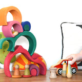 A child playing with a red Lanka Kade push along car with a Glückskäfer Gable House wooden stacking toy, stacked up in the background. Other wooden toys such as the Grapat Nins, wooden spools and a Holztiger oak tree have been added to the small world play scene set up. 