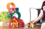 A child playing with a red Lanka Kade push along car with a Glückskäfer Gable House wooden stacking toy, stacked up in the background. Other wooden toys such as the Grapat Nins, wooden spools and a Holztiger oak tree have been added to the small world play scene set up. 
