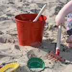 The Glückskäfer Red Metal Bucket placed on sand at a beach. A child's hand can be seen to the side using the red rake tool.