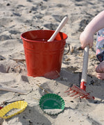 The Glückskäfer Red Metal Bucket placed on sand at a beach. A child's hand can be seen to the side using the red rake tool.