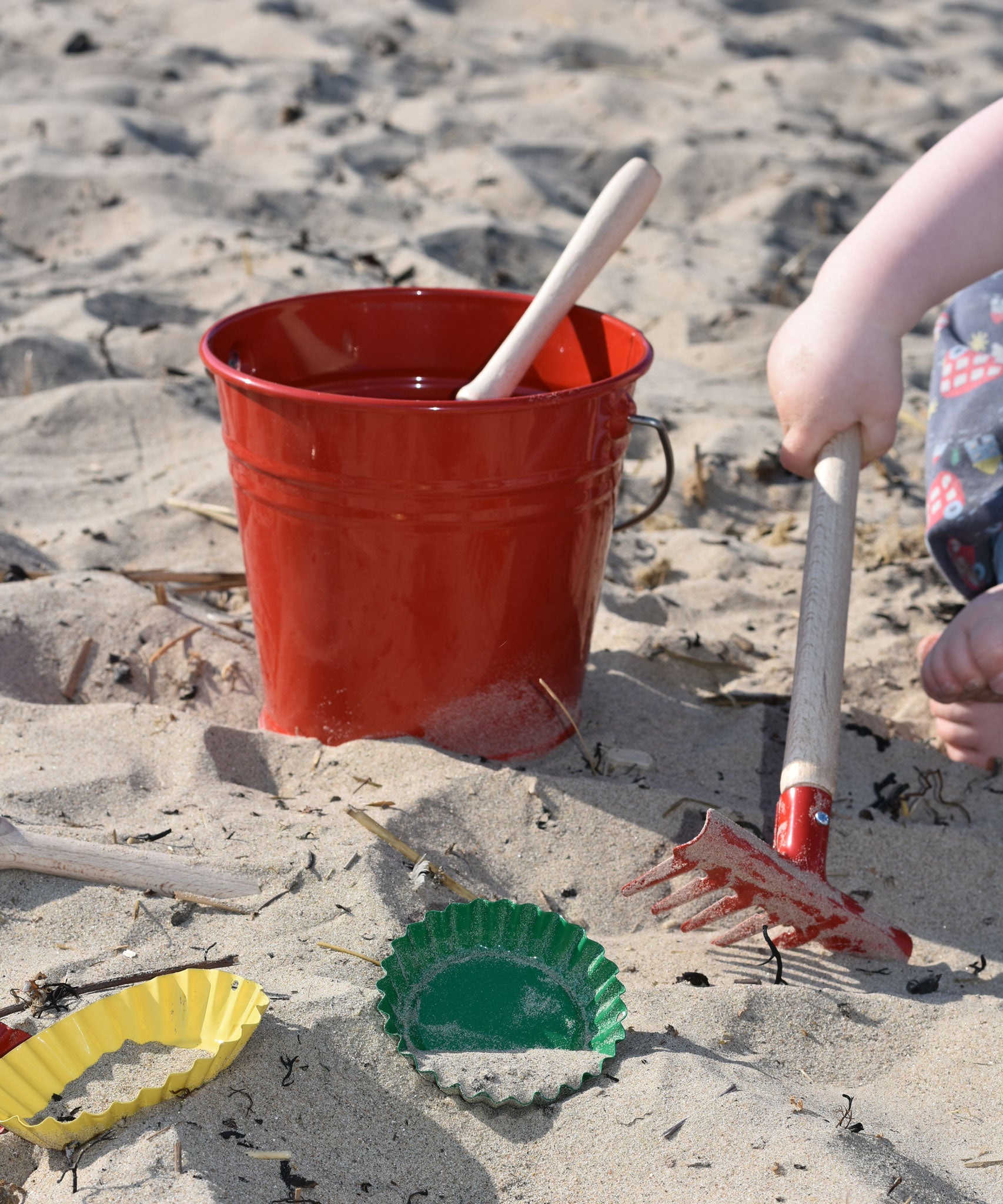 The Glückskäfer Red Metal Bucket placed on sand at a beach. A child's hand can be seen to the side using the red rake tool.