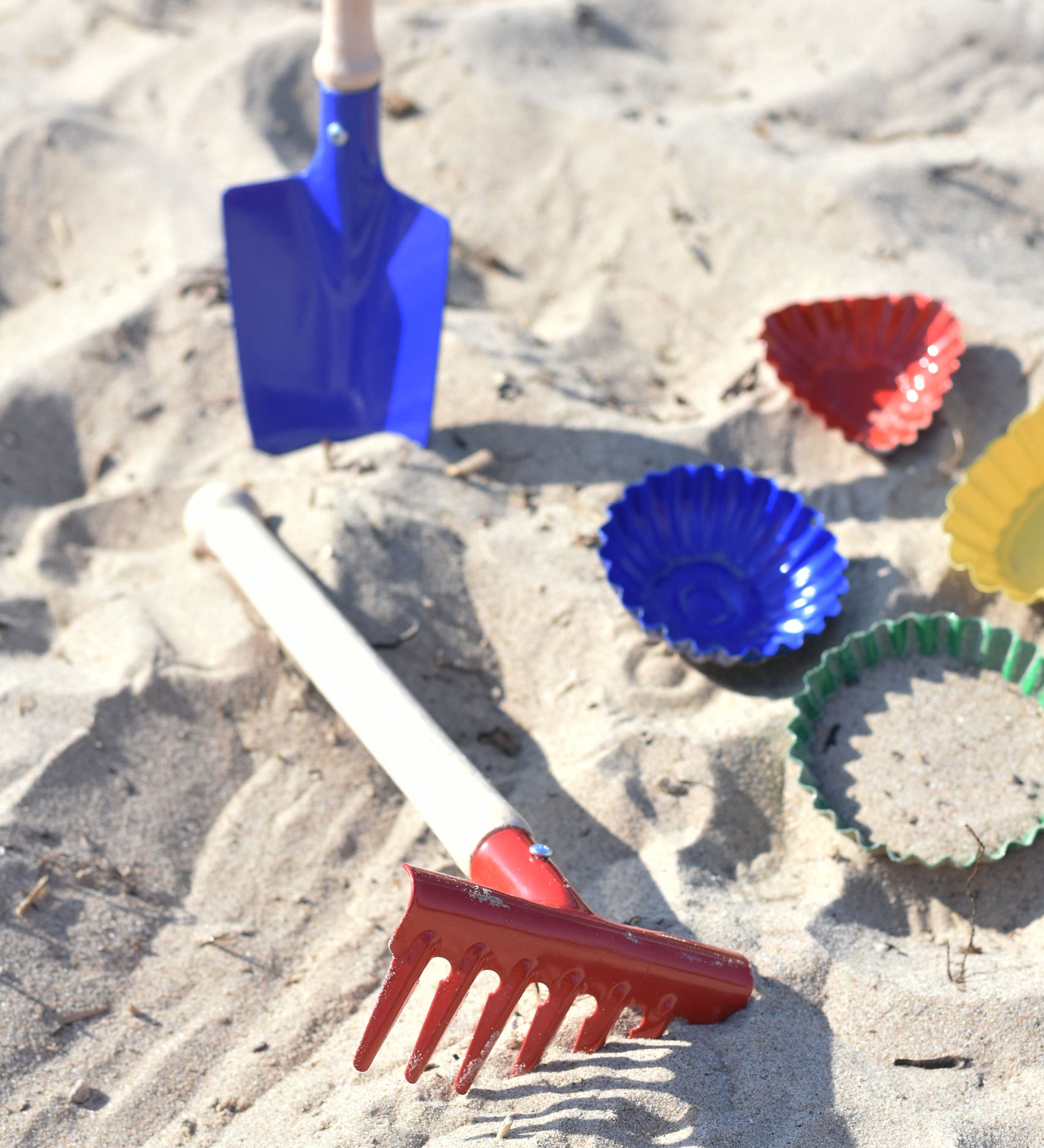 A Glückskäfer Metal Red Rake placed on the sand next to metal sand moulds. A blue round shovel can be seen in the background.