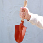 A child holding up a Gluckskafer red round metal shovel above a pot fill with soil.