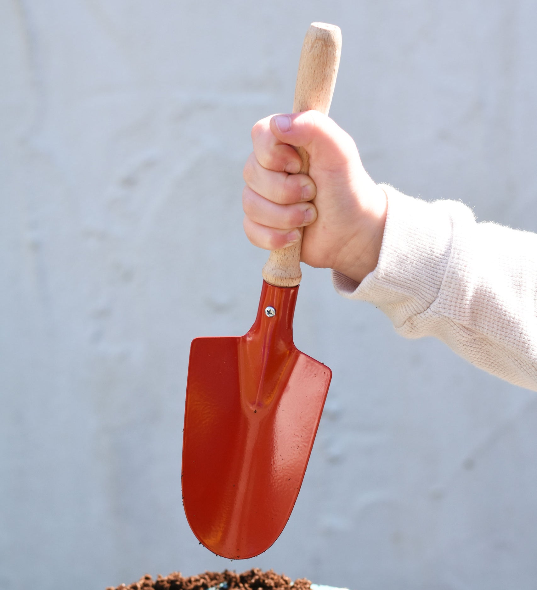A child holding up a Gluckskafer red round metal shovel above a pot fill with soil.