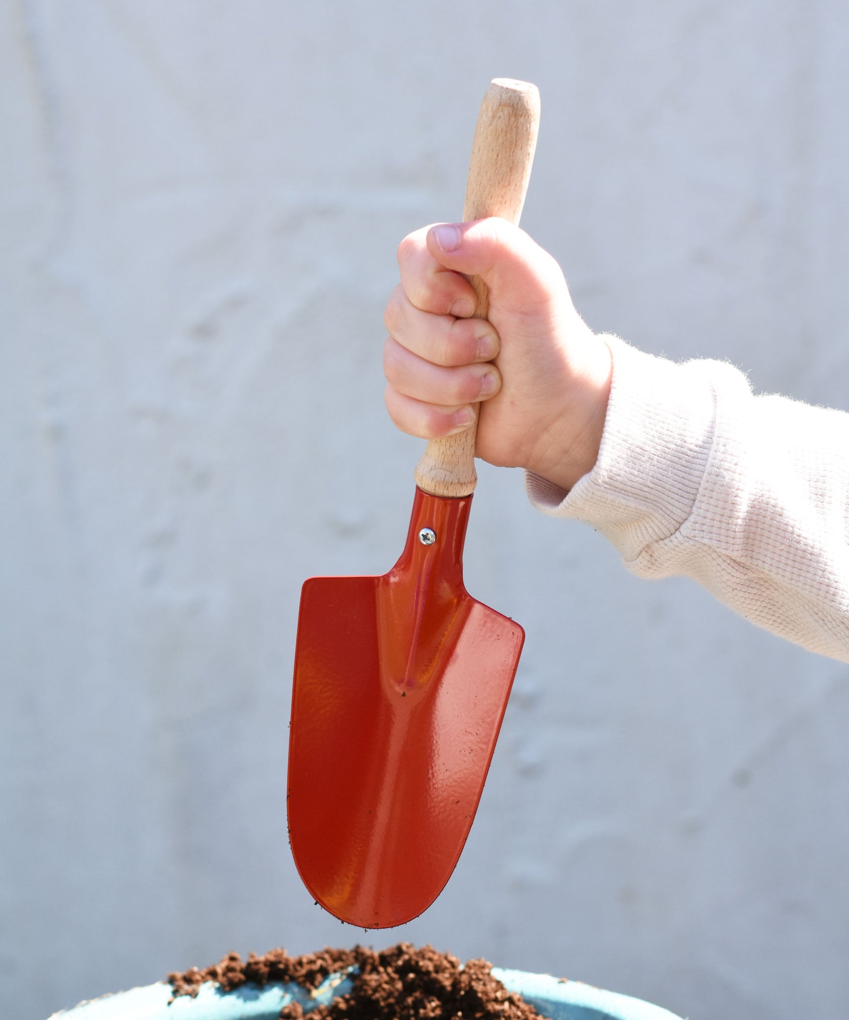 A child holding up a Gluckskafer red round metal shovel above a pot fill with soil.