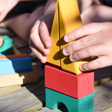 A close up of a child's hand placing the two yellow triangular shaped wooden building blocks from the Glückskäfer 20-Piece Small Toscana Blocks Set on top of a small stack. 