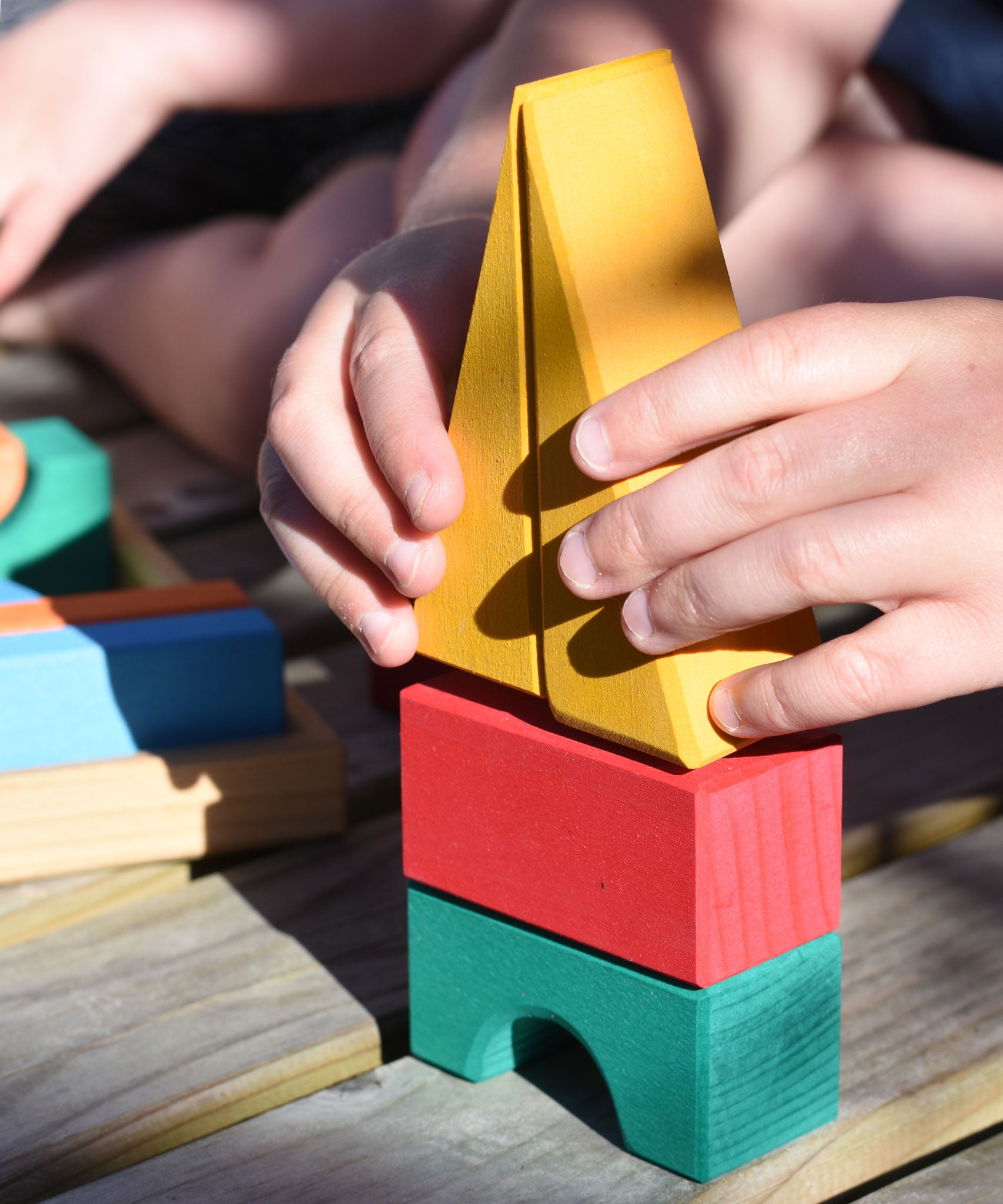A close up of a child's hand placing the two yellow triangular shaped wooden building blocks from the Glückskäfer 20-Piece Small Toscana Blocks Set on top of a small stack. 