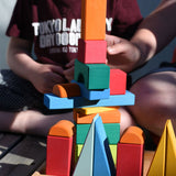 A close up of two children sitting on wooden decking outdoors playing with the Glückskäfer 20-Piece Small Toscana Blocks Set. They are stacking the blocks on the wooden tray. 