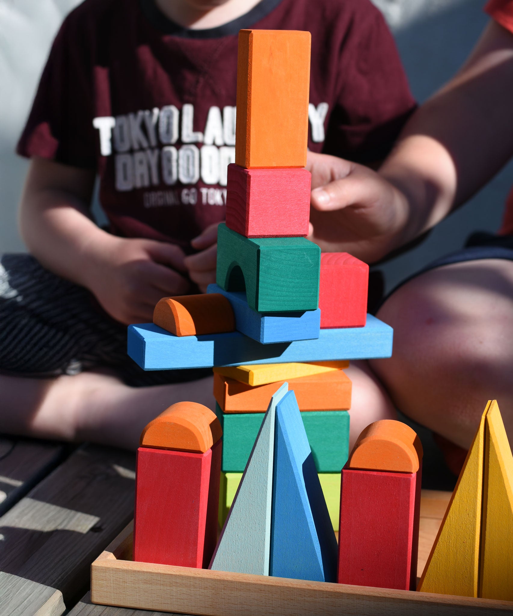 A close up of two children sitting on wooden decking outdoors playing with the Glückskäfer 20-Piece Small Toscana Blocks Set. They are stacking the blocks on the wooden tray. 