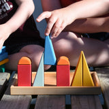 Children playing with the Glückskäfer 20-Piece Small Toscana Blocks outdoors on wooden decking. A child can be seen placing a blue coloured triangular shaped block onto the wooden tray in a line up of other blocks from the set.
