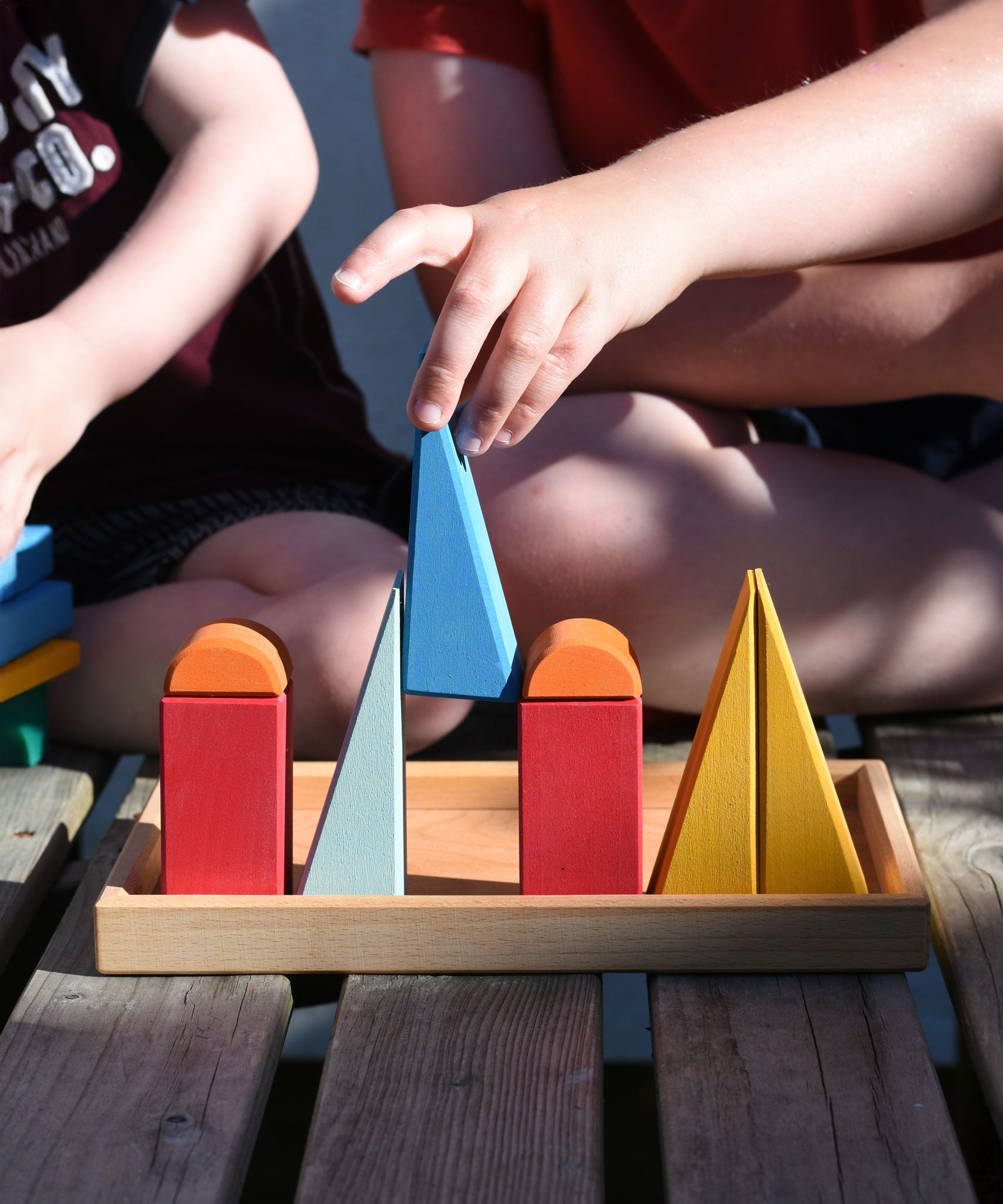 Children playing with the Glückskäfer 20-Piece Small Toscana Blocks outdoors on wooden decking. A child can be seen placing a blue coloured triangular shaped block onto the wooden tray in a line up of other blocks from the set.