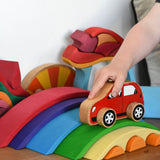 The Gluckskafer Rainbow Bridge wooden toy which consists of 5 arch shaped bridge pieces and 3 small semi circle pieces than fit underneath the smallest bridge piece. A child's hand can be seen playing with a red Lanka Kade toy car, driving it over the green coloured bridge piece. 
