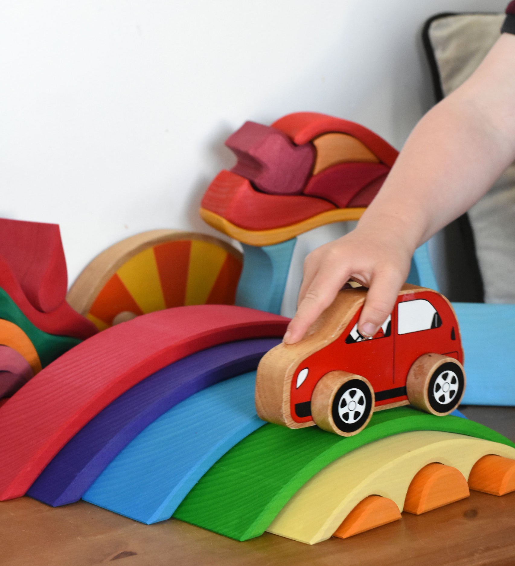 The Gluckskafer Rainbow Bridge wooden toy which consists of 5 arch shaped bridge pieces and 3 small semi circle pieces than fit underneath the smallest bridge piece. A child's hand can be seen playing with a red Lanka Kade toy car, driving it over the green coloured bridge piece. 

