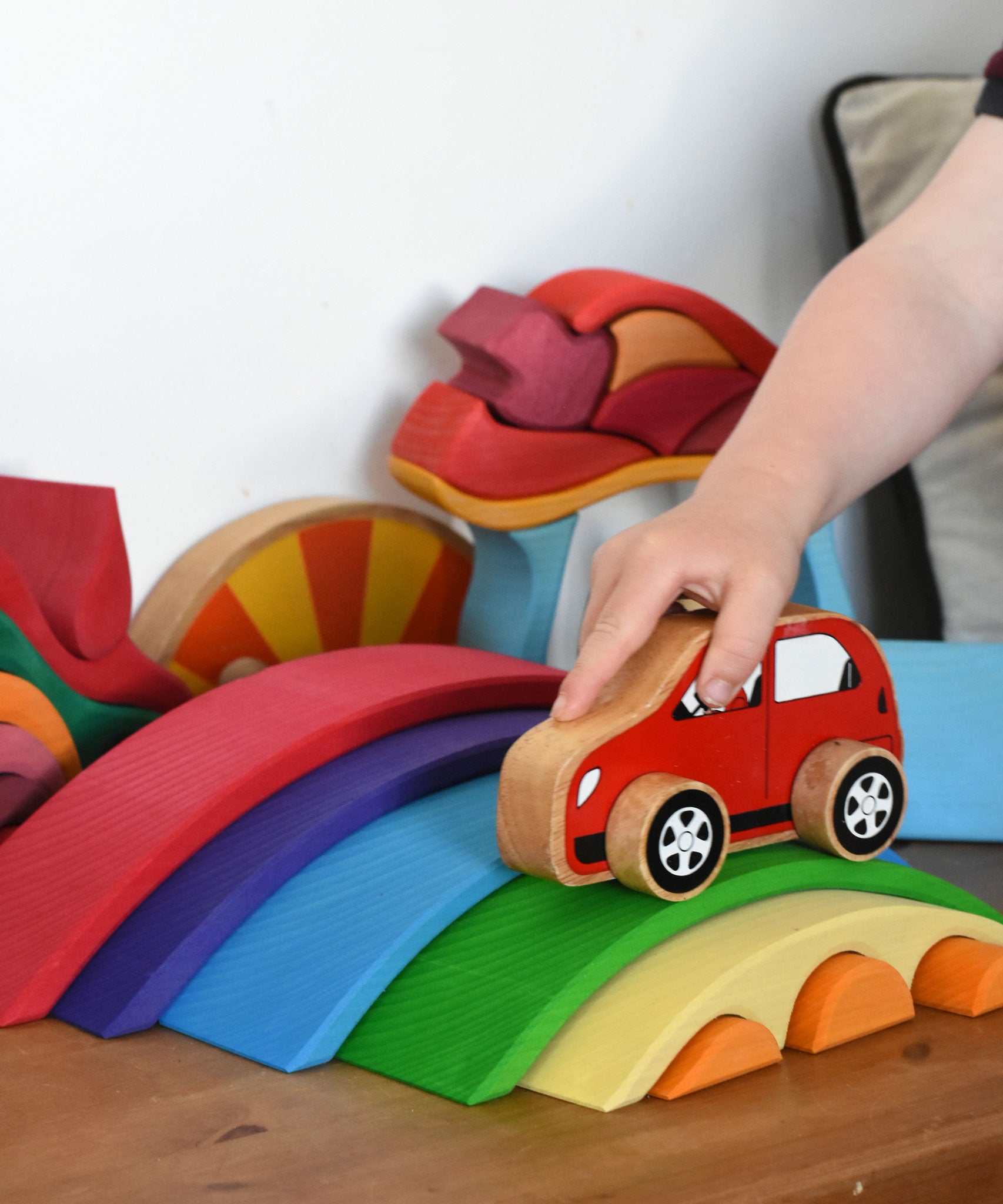 The Gluckskafer Rainbow Bridge wooden toy which consists of 5 arch shaped bridge pieces and 3 small semi circle pieces than fit underneath the smallest bridge piece. A child's hand can be seen playing with a red Lanka Kade toy car, driving it over the green coloured bridge piece. 
