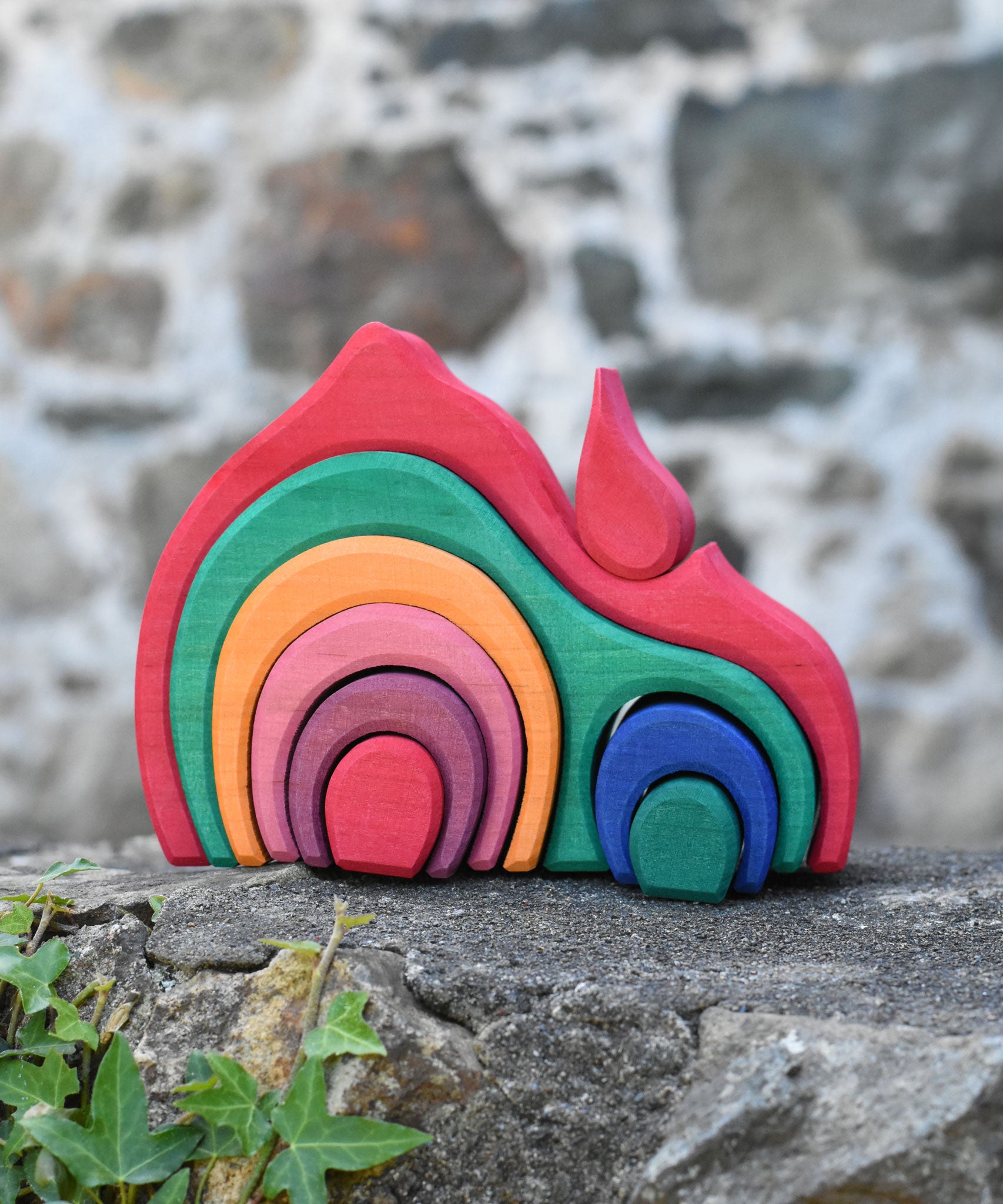 The Glückskäfer Gable House wooden toy placed on a stone wall outdoors. The set has 9 wooden pieces that fit together snuggly to form the complete red gable house.