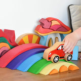 A close up of a child playing with the Gluckskafer Rainbow Bridge. The child is placing a red coloured wooden Lanka Kade car on thegreen coloured bridge piece from the Gluckskafer Rainbow Bridge. Other wooden toys can be seen in the background. 
