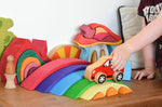 A close up of a child playing with the Gluckskafer Rainbow Bridge. The child is placing a red coloured wooden Lanka Kade car on thegreen coloured bridge piece from the Gluckskafer Rainbow Bridge. Other wooden toys can be seen in the background. 

