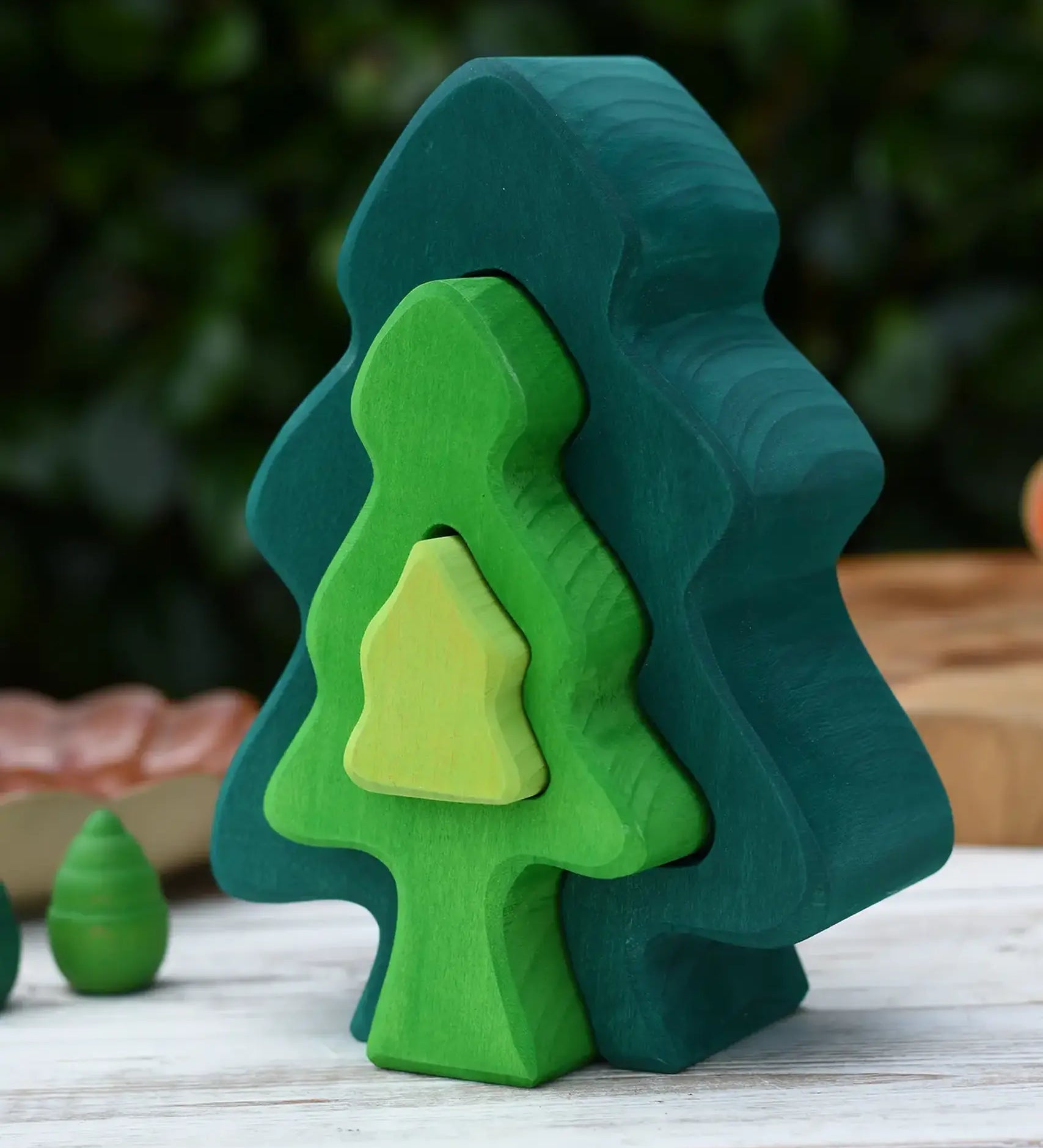 Glückskäfer 6-Piece Fir Tree wooden tree stacking toy with six nested tree shapes in shades of green placed on a wooden surface outdoors.