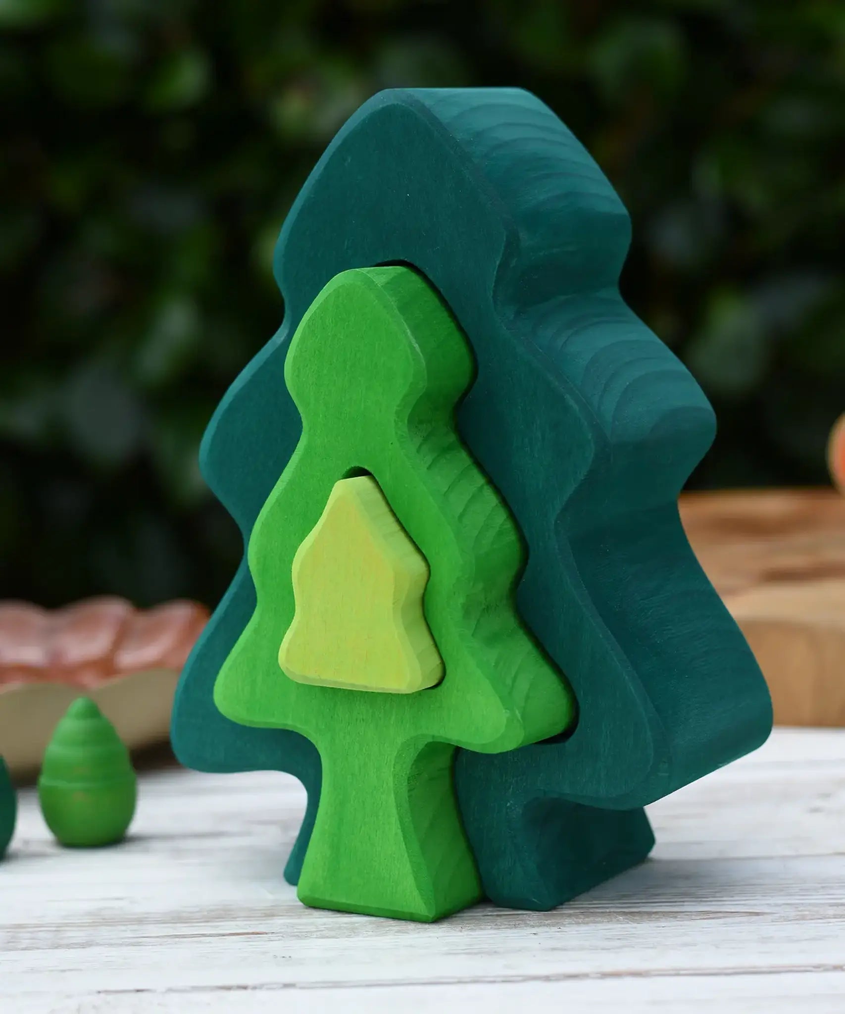 Glückskäfer 6-Piece Fir Tree wooden tree stacking toy with six nested tree shapes in shades of green placed on a wooden surface outdoors.