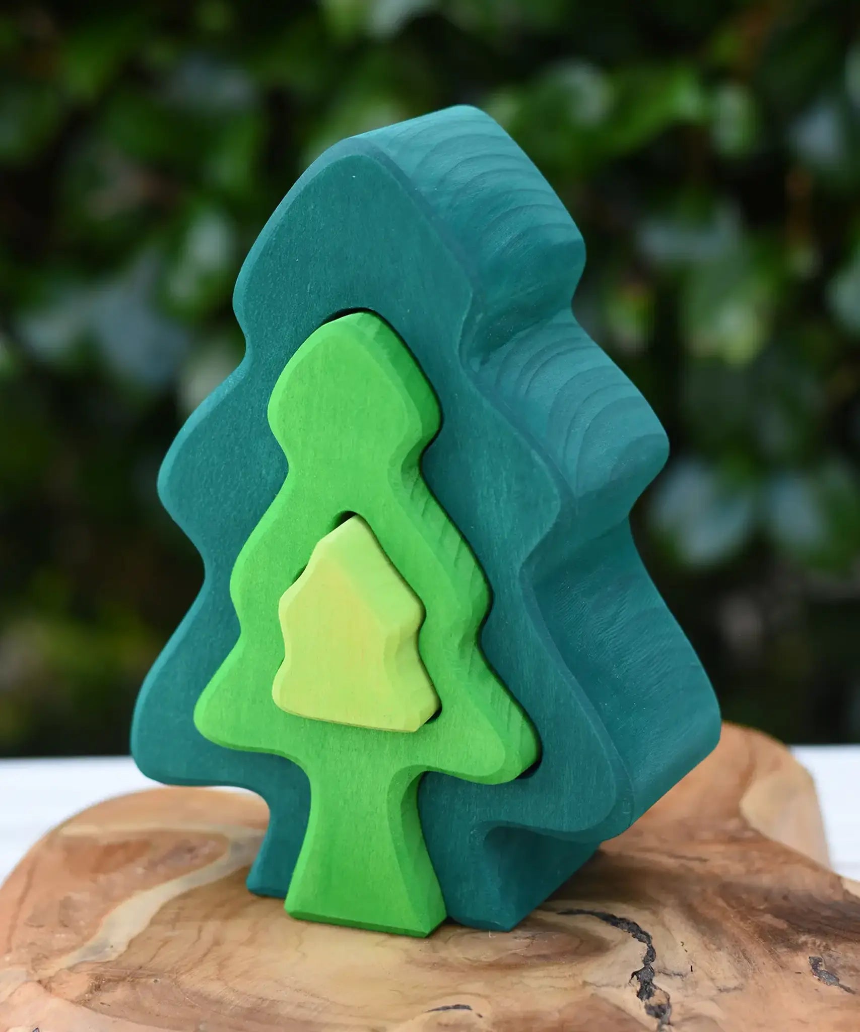 Glückskäfer 6-Piece Fir Tree wooden tree stacking toy with six nested tree shapes in shades of green placed on a wooden surface outdoors.