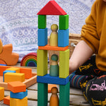 A tower shaped stack created with blocks from the Glückskäfer 29-Piece House Blocks set. A child can be sitting to one side and other wooden toys including a mini Grimm's rainbow can be seen in the background.