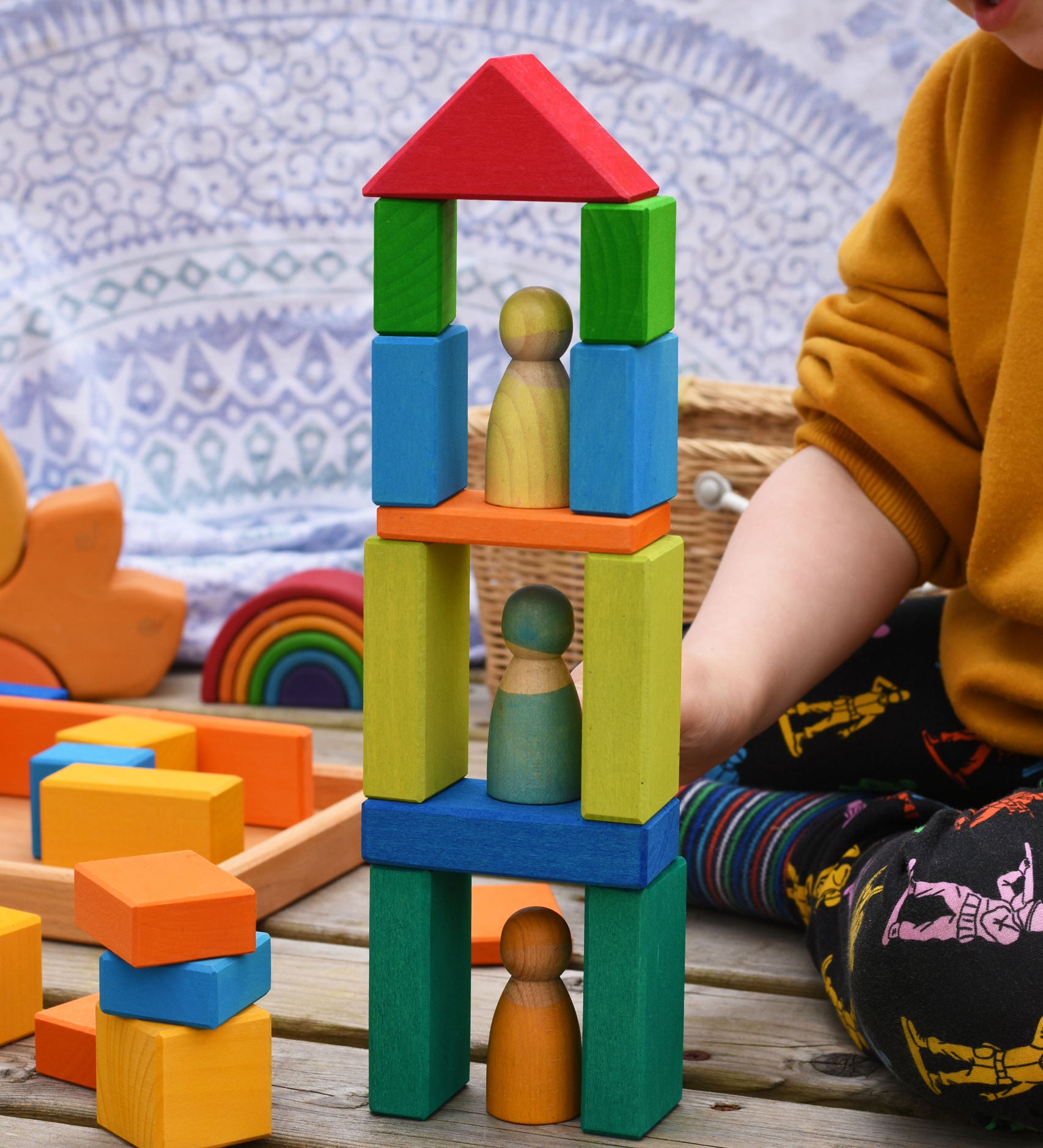 A tower shaped stack created with blocks from the Glückskäfer 29-Piece House Blocks set. A child can be sitting to one side and other wooden toys including a mini Grimm's rainbow can be seen in the background.