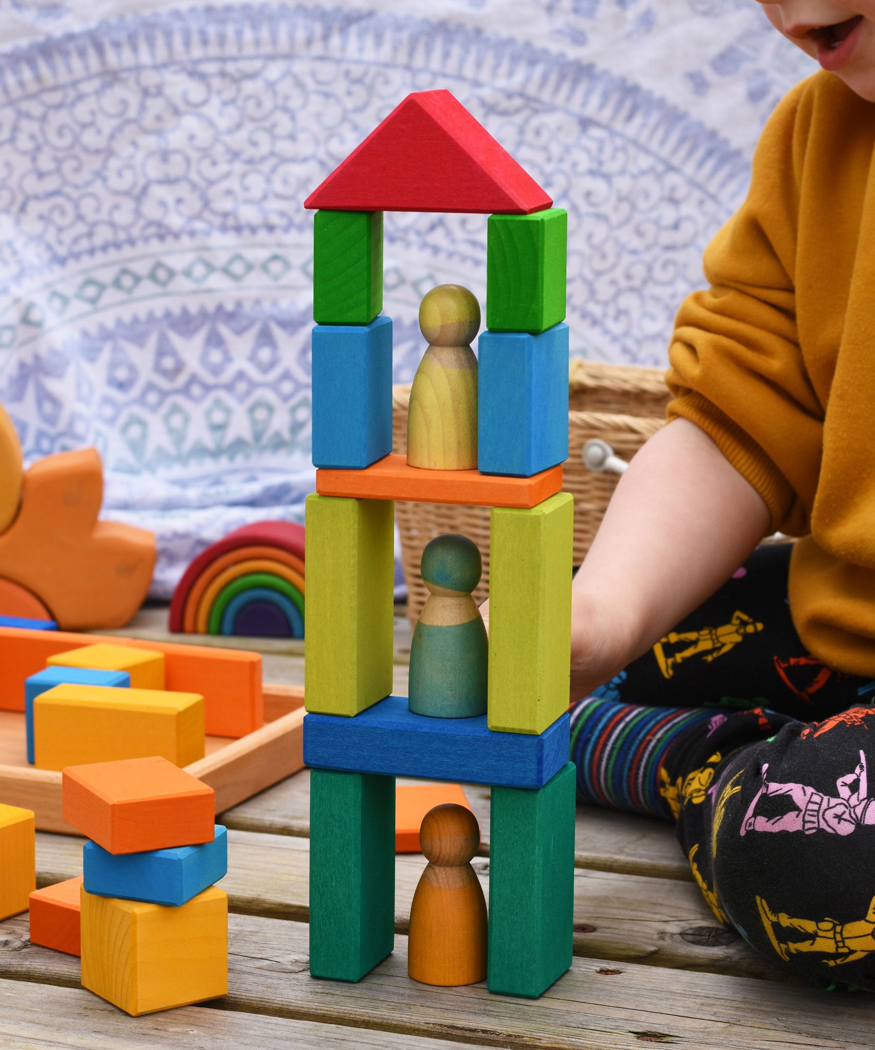A tower shaped stack created with blocks from the Glückskäfer 29-Piece House Blocks set. A child can be sitting to one side and other wooden toys including a mini Grimm's rainbow can be seen in the background.