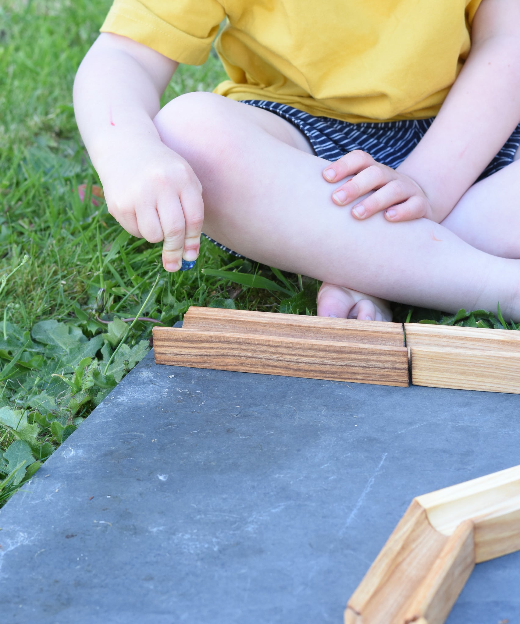A child placing a glass marble on the Glückskäfer 23 Piece Wooden Marble Track laid out onto a slate surface outdoors. 