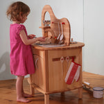A young child playing at the Gluckskafer wooden play kitchen island, using the sink and utensils during pretend play.