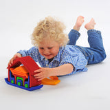 A child laying on his front playing with the Glückskäfer All-in Rainbow coloured House. The child has taken some pieces out of the house and is lifting the large red coloured house shaped piece from the base.