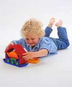 A child laying on his front playing with the Glückskäfer All-in Rainbow coloured House. The child has taken some pieces out of the house and is lifting the large red coloured house shaped piece from the base.