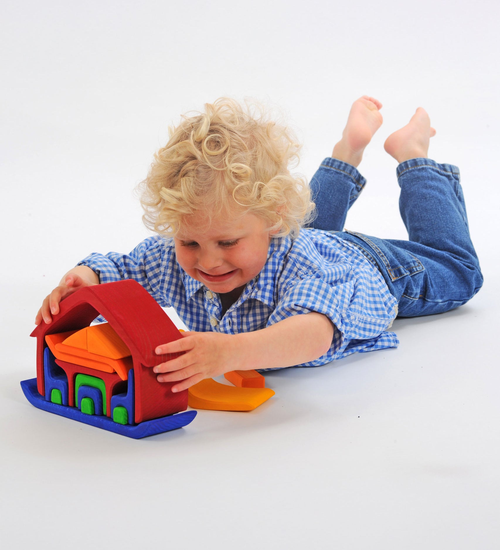 A child laying on his front playing with the Glückskäfer All-in Rainbow coloured House. The child has taken some pieces out of the house and is lifting the large red coloured house shaped piece from the base.