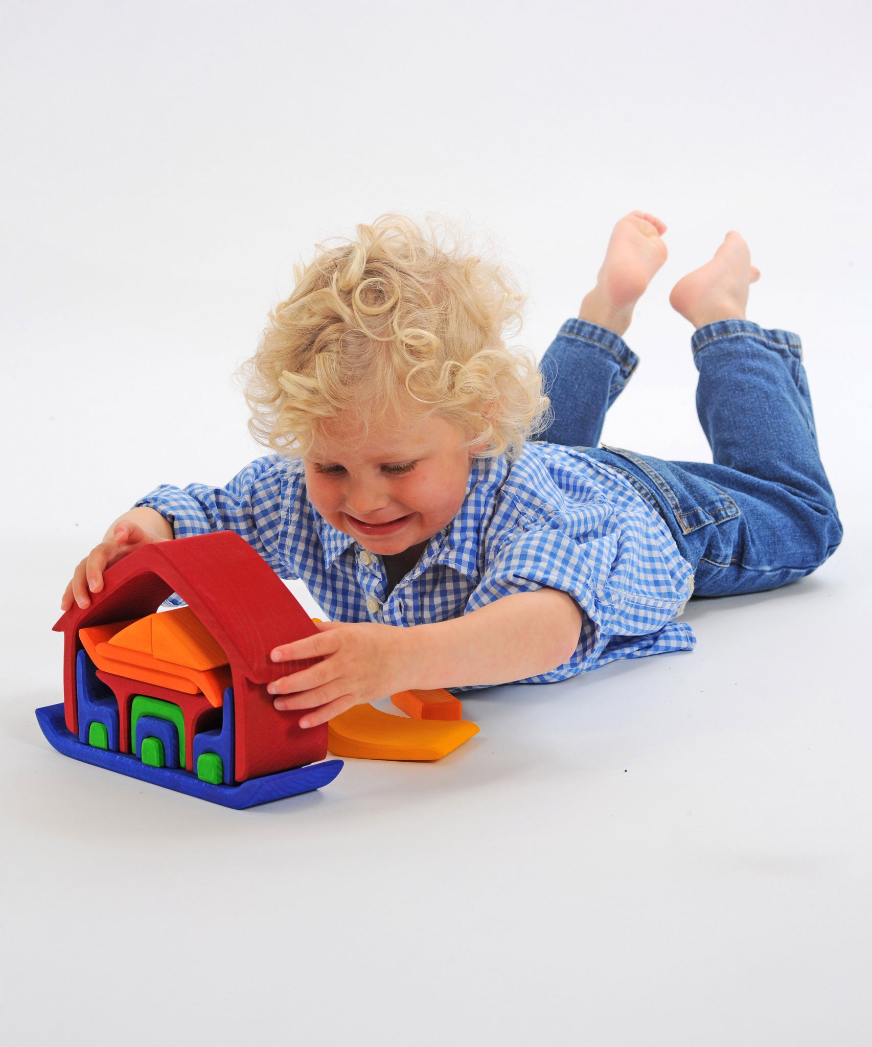 A child laying on his front playing with the Glückskäfer All-in Rainbow coloured House. The child has taken some pieces out of the house and is lifting the large red coloured house shaped piece from the base.