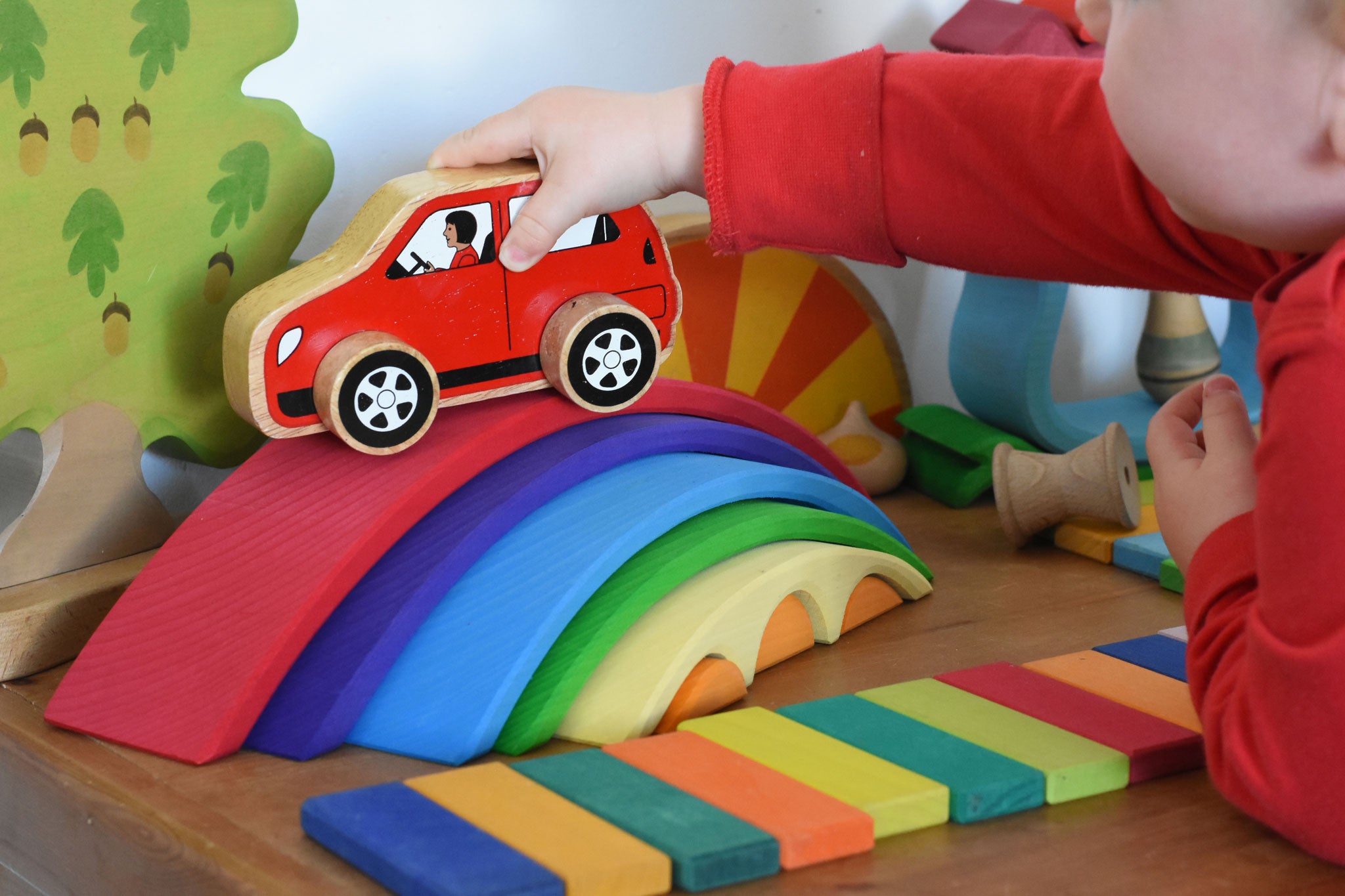 A close up of a child playing with the Gluckskafer Rainbow Bridge. The child is placing a red coloured wooden Lanka Kade car on the red coloured largest bridge piece from the Gluckskafer Rainbow Bridge. The Gluckskafer wooden slats can be seen laid out in front of the Gluckskafer Rainbow Bridge with other wooden toy in the background. 
