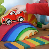 A close up of a child playing with the Gluckskafer Rainbow Bridge. The child is placing a red coloured wooden Lanka Kade car on the red coloured largest bridge piece from the Gluckskafer Rainbow Bridge. The Gluckskafer wooden slats can be seen laid out in front of the Gluckskafer Rainbow Bridge with other wooden toy in the background. 
