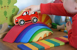 A close up of a child playing with the Gluckskafer Rainbow Bridge. The child is placing a red coloured wooden Lanka Kade car on the red coloured largest bridge piece from the Gluckskafer Rainbow Bridge. The Gluckskafer wooden slats can be seen laid out in front of the Gluckskafer Rainbow Bridge with other wooden toy in the background. 
