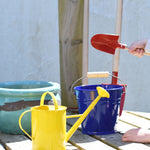 A yellow Glückskäfer Children's 1L Metal Watering Can placed on wooden decking. A blue bucket and other children's gardening tools can be seen in the background.