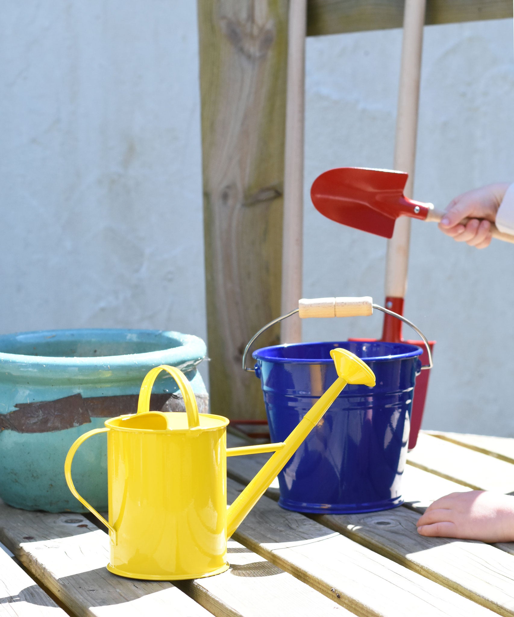A yellow Glückskäfer Children's 1L Metal Watering Can placed on wooden decking. A blue bucket and other children's gardening tools can be seen in the background.