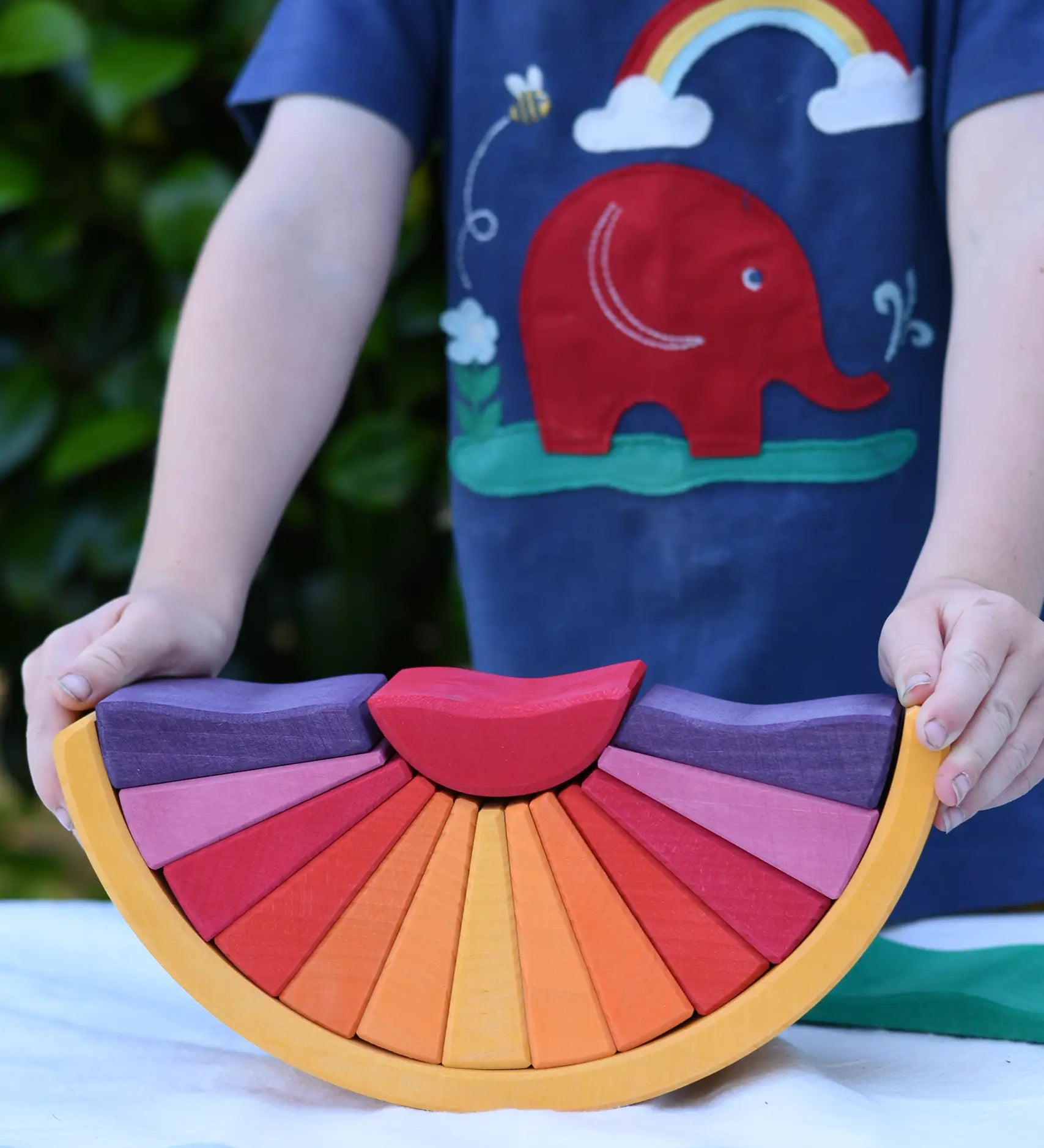 A child holding the Glückskäfer wooden stacking sunray arch in yellow purple placed on a table outdoors. 