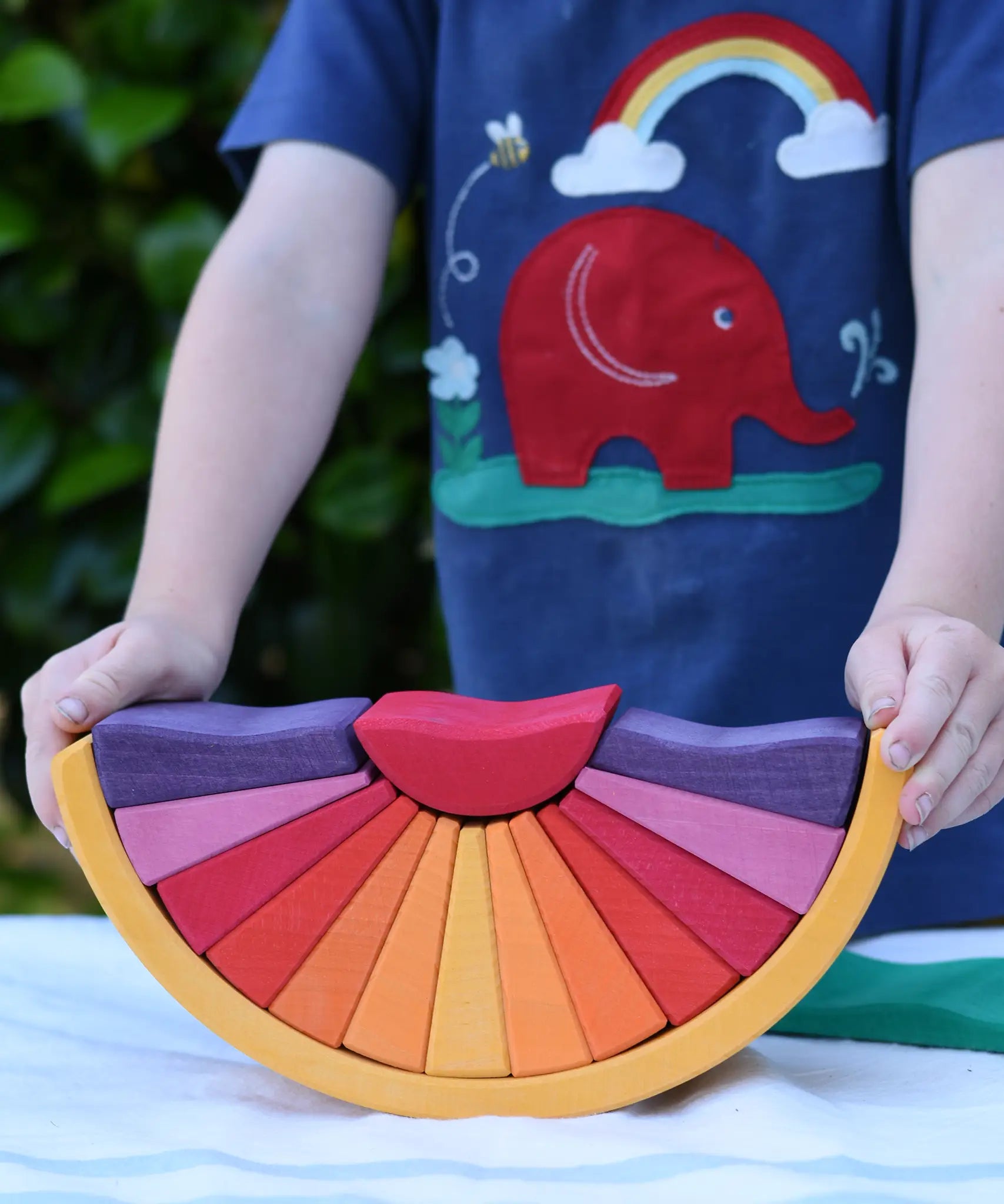 A child holding the Glückskäfer wooden stacking sunray arch in yellow purple placed on a table outdoors. 
